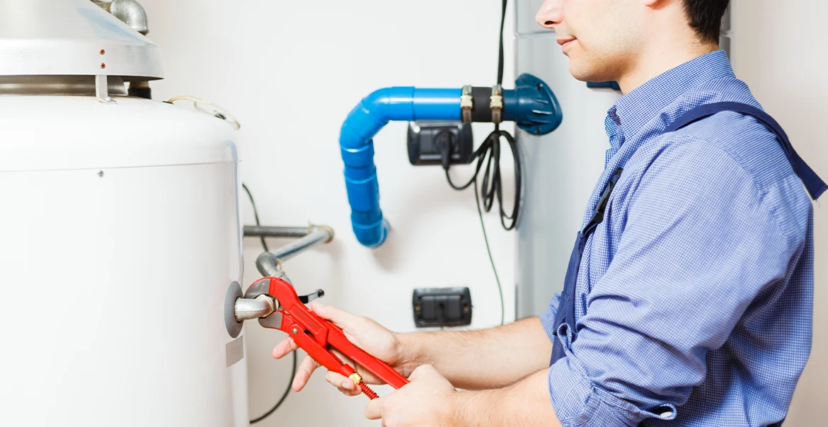 HVAC Technician using a red wrench to work on a water heater beside blue plumbing pipes