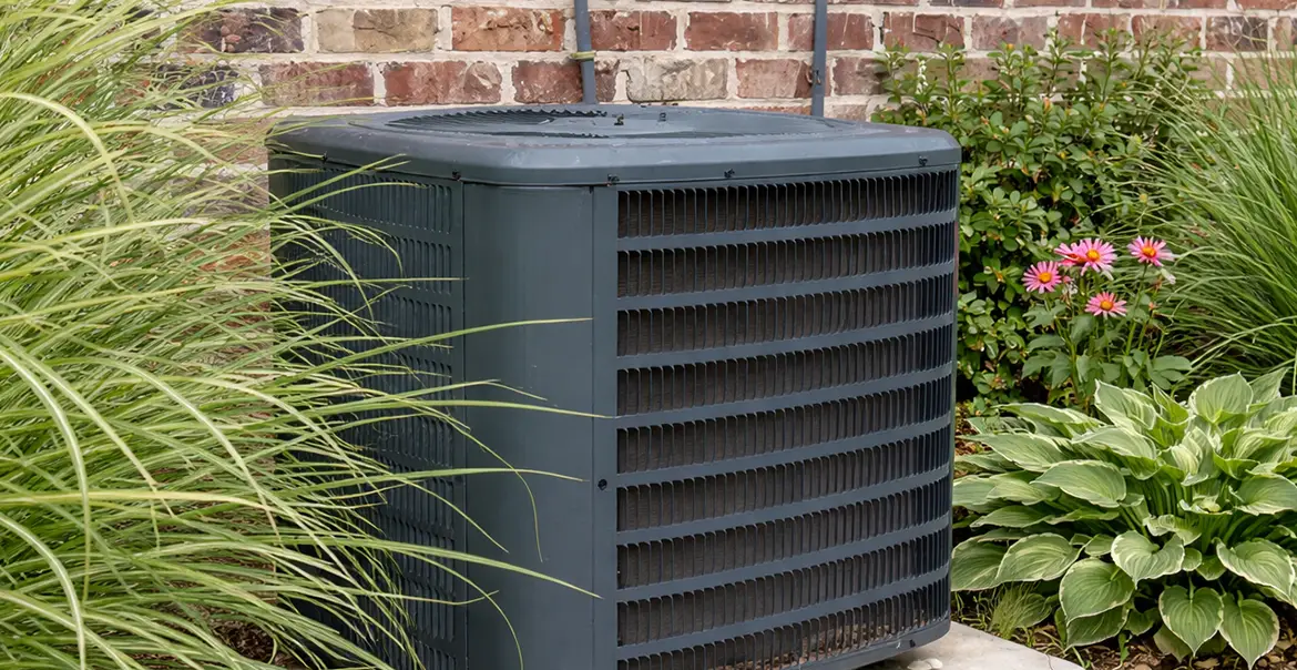 Outdoor heat pump unit installed beside a brick house surrounded by plants